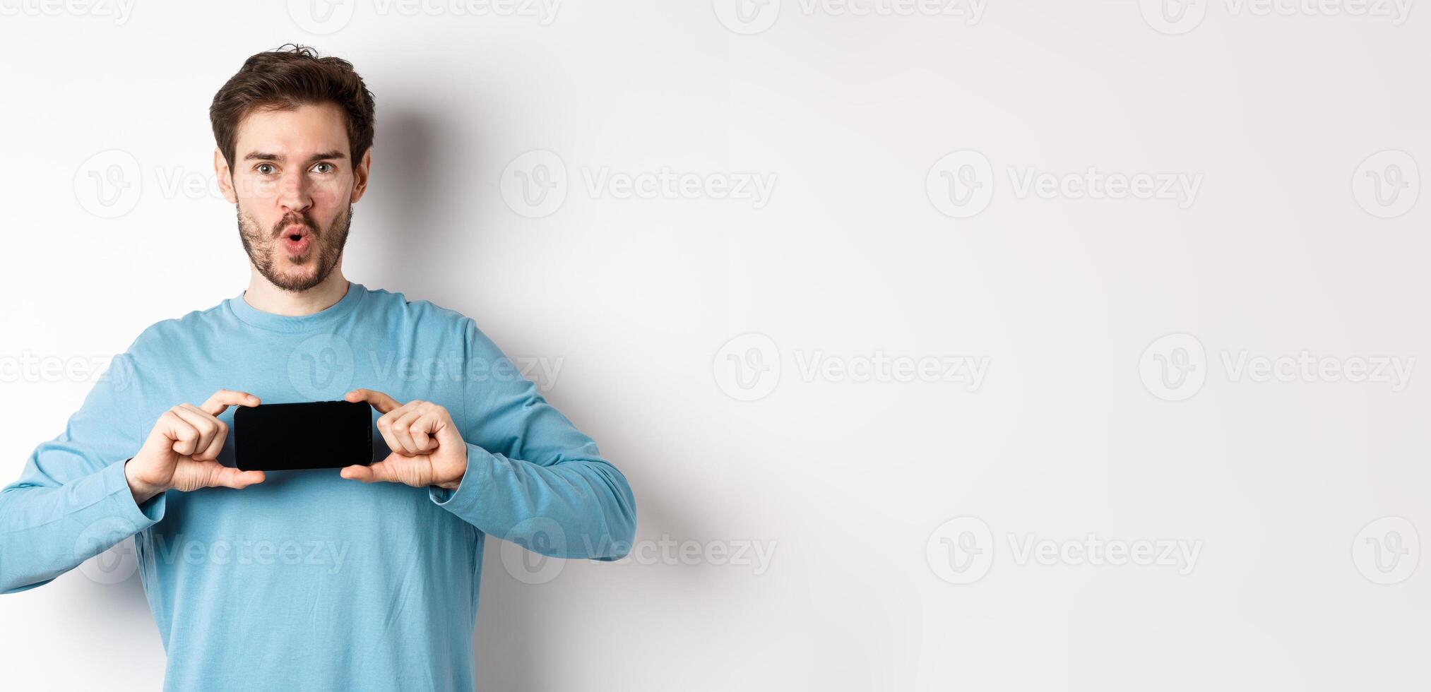 E-commerce and shopping concept. Image of impressed man saying wow and showing blank mobile phone screen in horizontal, standing over white background photo
