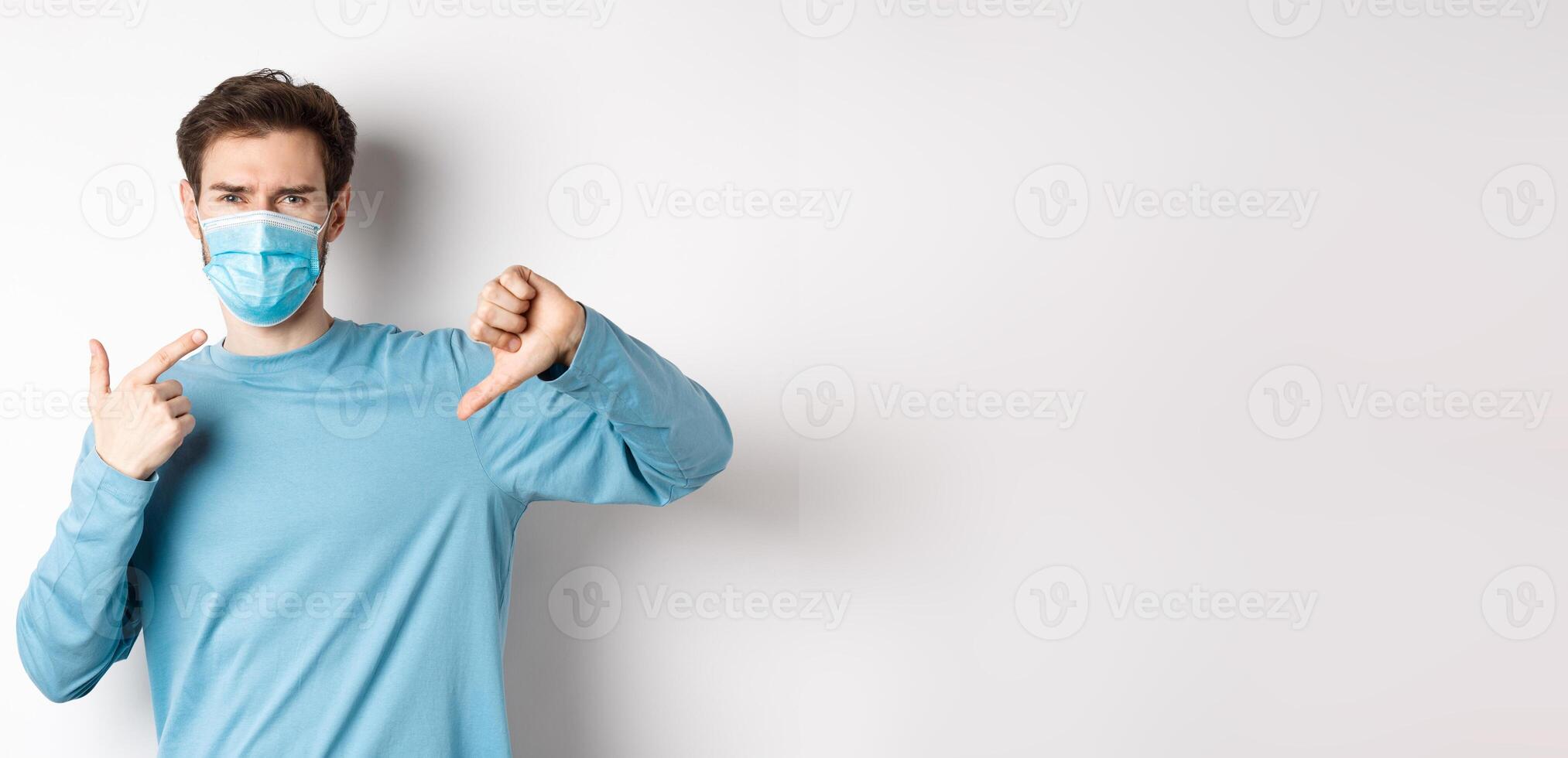 Coronavirus, health and quarantine concept. Angry man pointing at his medical mask and showing thumbs down, complaining on covid, white background photo