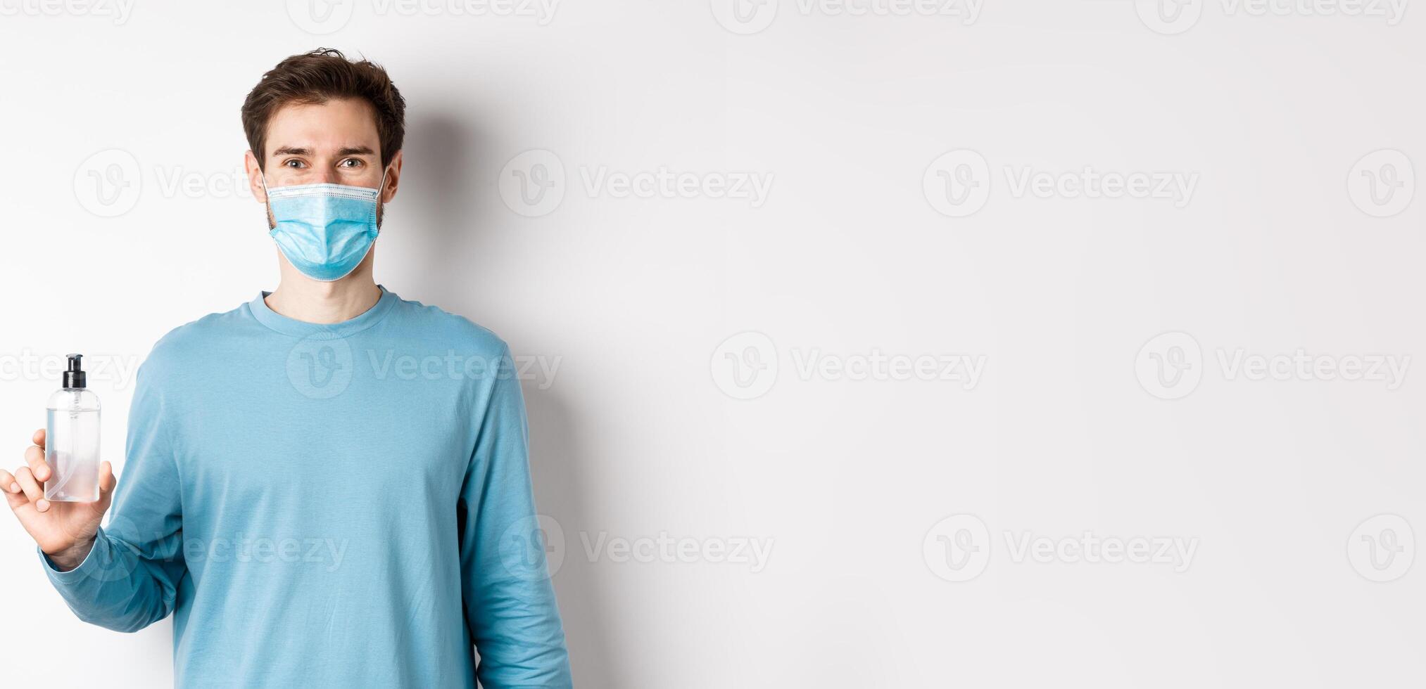Covid-19, health and quarantine concept. Portrait of smiling man in medical mask showing hand sanitizer bottle, standing over white background photo