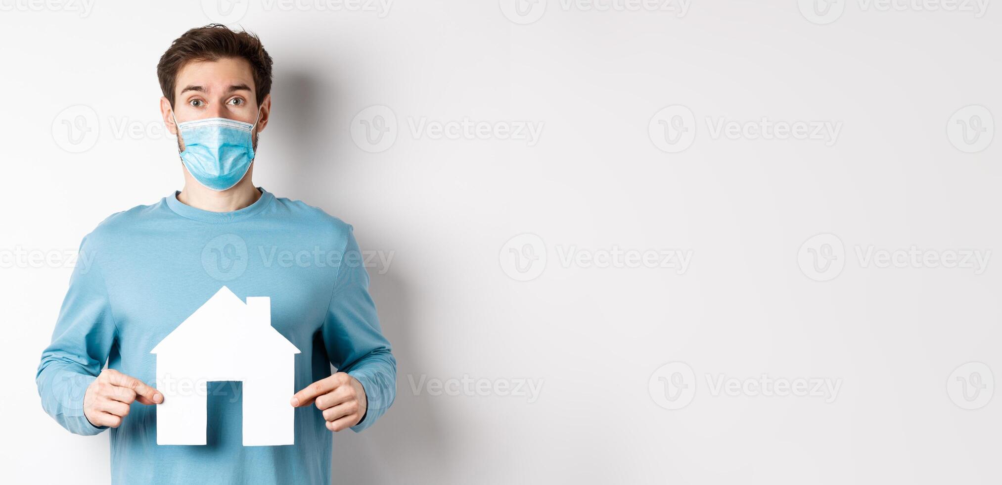 Covid and real estate concept. Surprised man in medical mask showing paper house cutout, looking at camera, white background photo