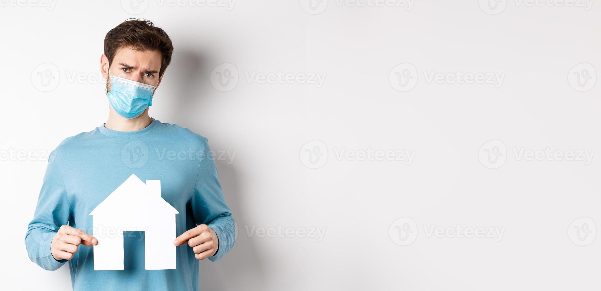 Covid and real estate concept. Sad and doubtful young man in medical mask feeling reluctant, showing paper home cutout, standing over white background photo