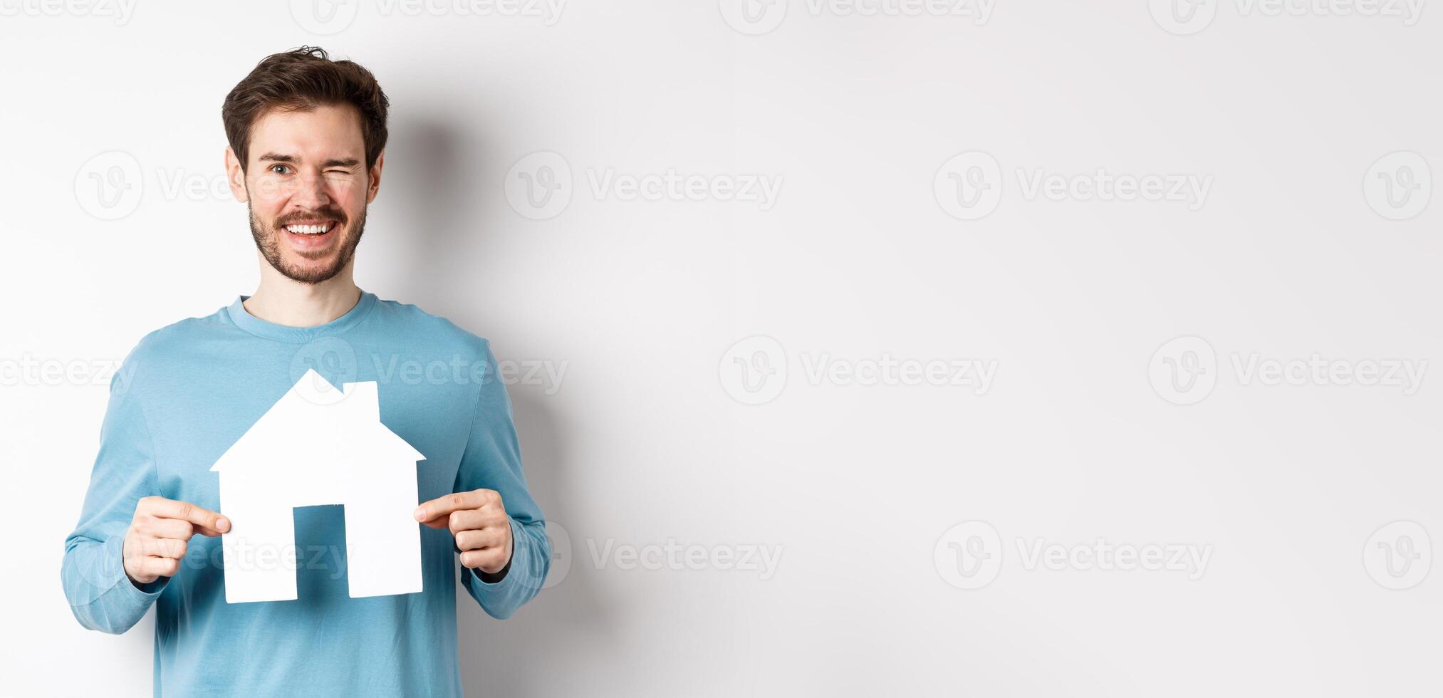 Real estate and insurance concept. Happy young man winking and smiling, showing paper house cutout, standing over white background photo