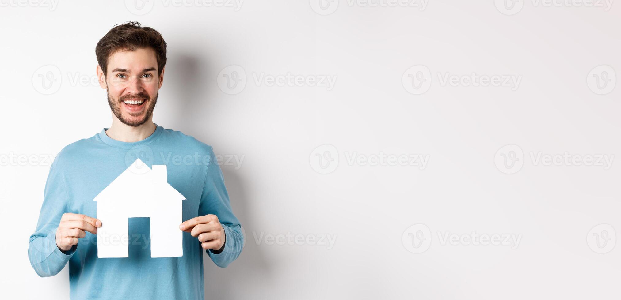 Real estate and insurance concept. Handsome modern man buying property, smiling and showing paper house cutout, standing over white background photo