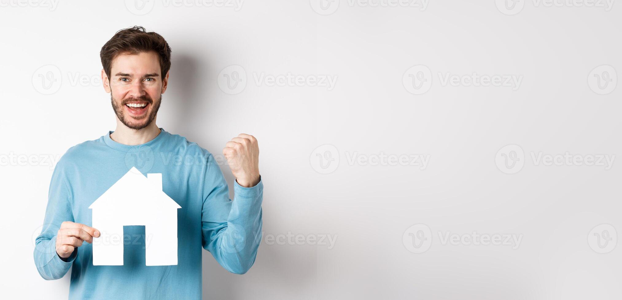 Real estate and insurance concept. Cheerful man buying property and celebrating, saying yes and showing paper house cutout, standing on white background photo