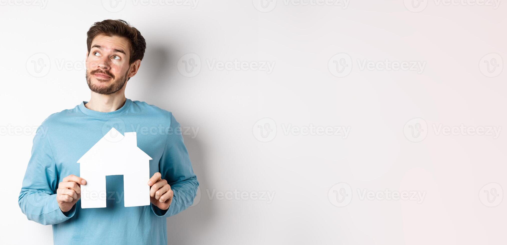 Real estate and insurance concept. Man dreaming about buying property, holding paper house cutout and looking at upper left corner, imaging things, white background photo