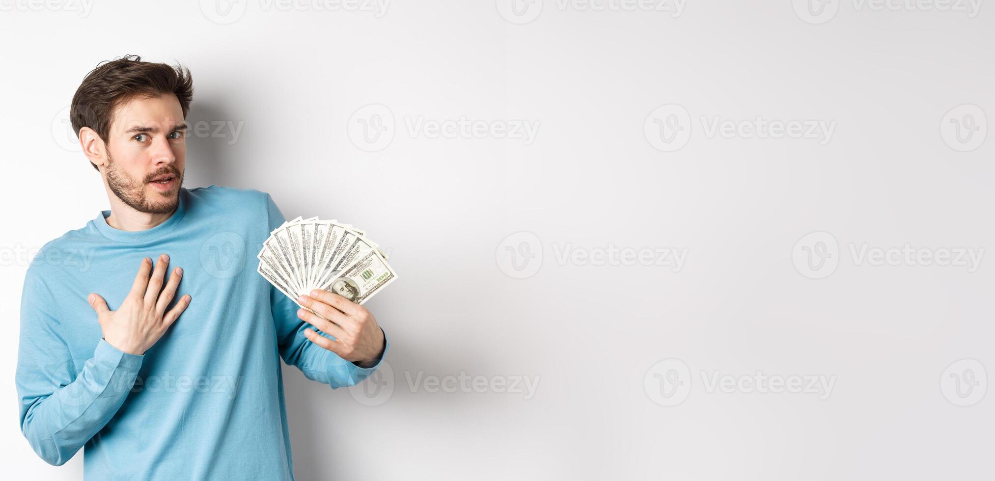 Surprised man holding money, looking amazed at camera, touching chest and gasping, standing in blue sweatshirt over white background photo