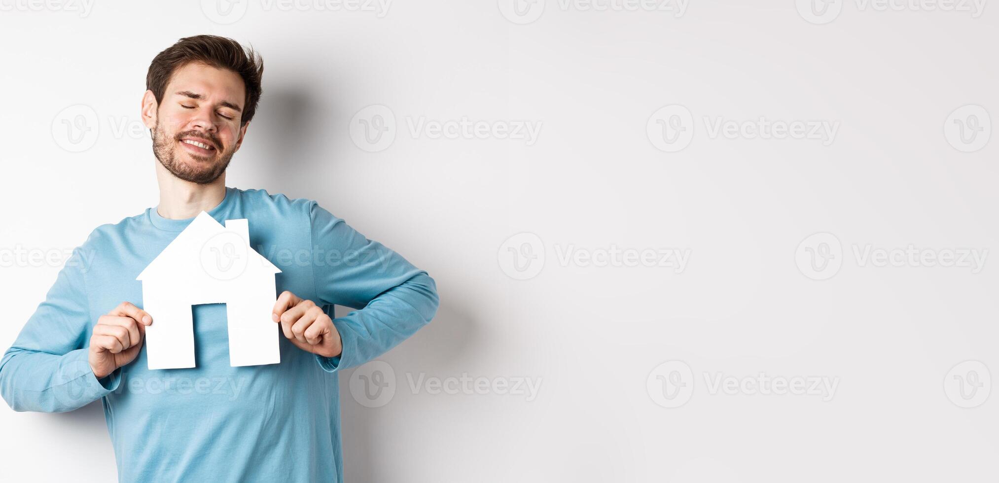Real estate and insurance concept. Dreamy young man smiling with closed eyes, showing paper house cutout, wishing to buy home, standing over white background photo