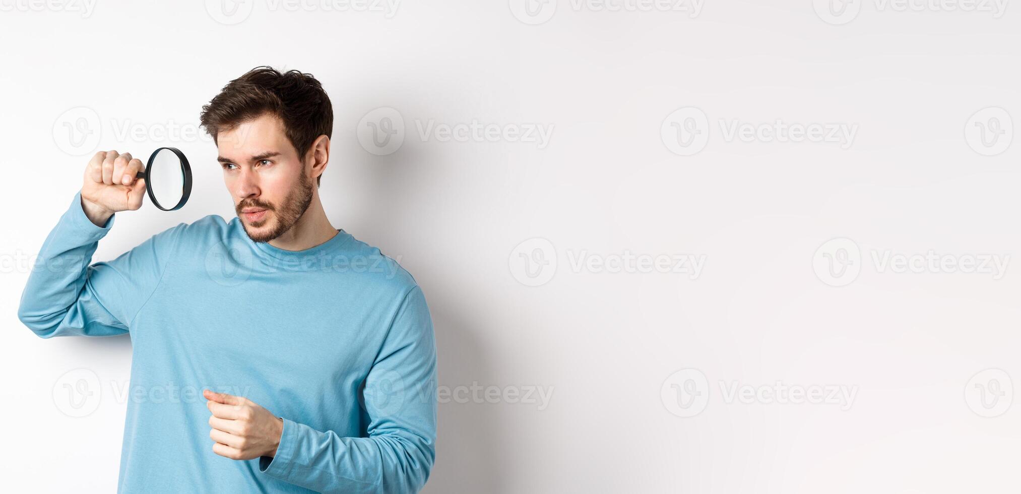 Young handsome man look through magnifying glass with curious face, investigating or searching for something, standing on white background photo