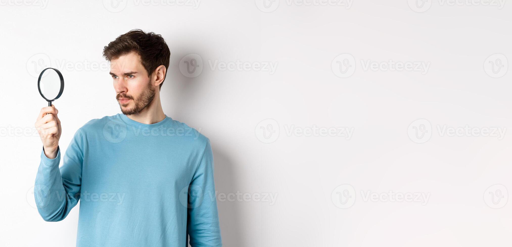 Young handsome man look through magnifying glass with curious face, investigating or searching for something, standing on white background photo