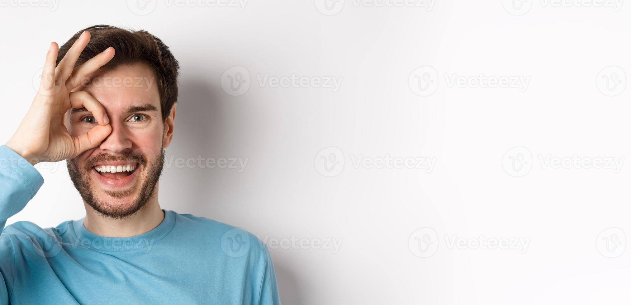 Excited smiling man looking through okay sign with happy face, approve and like promo offer, standing over white background photo