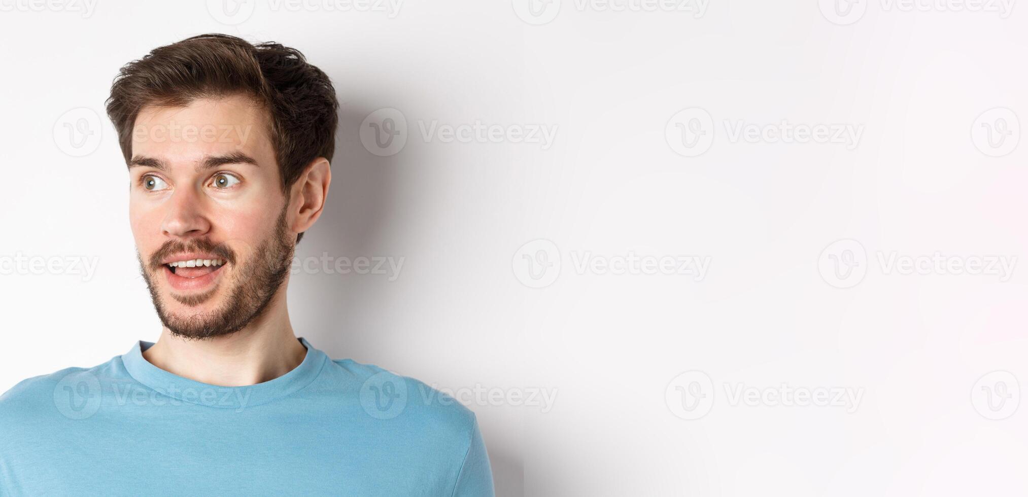 Close-up of surprised and excited bearded man, looking left with opened mouth and amazed face, checking out promo, white background photo