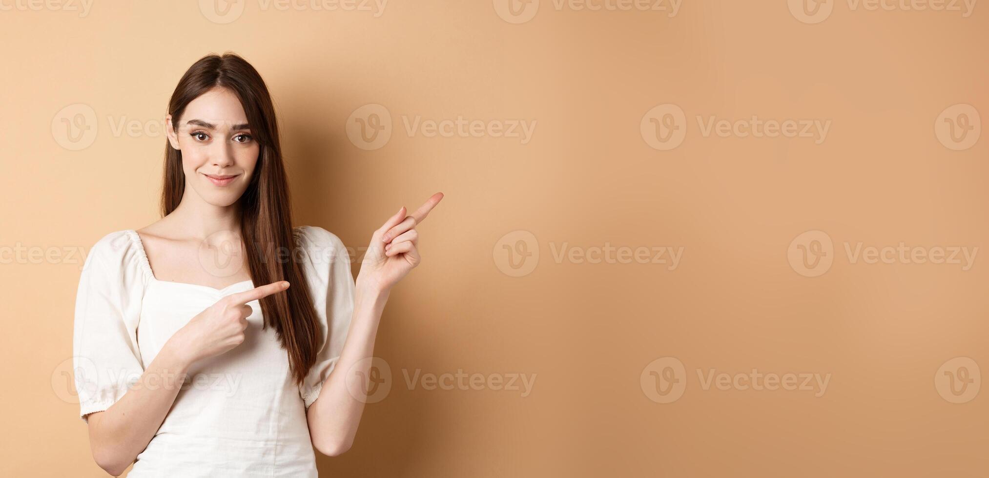 Beautiful young woman in dress pointing fingers right, showing logo and looking tender at camera, beige background photo