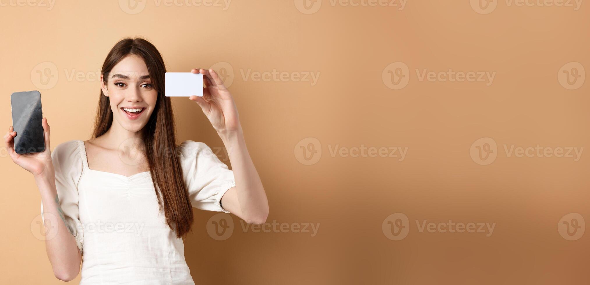 Cheerful young woman showing plastic credit card and empty mobile phone screen, smiling pleased at camera, standing on beige background photo