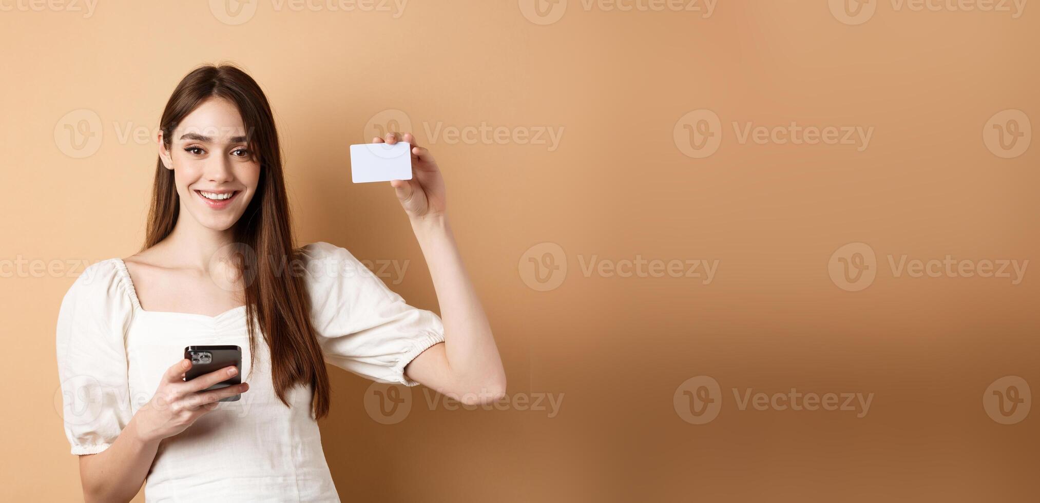 E-commerce concept. Smiling woman showing plastic credit card while shopping online on mobile phone, standing against beige background photo