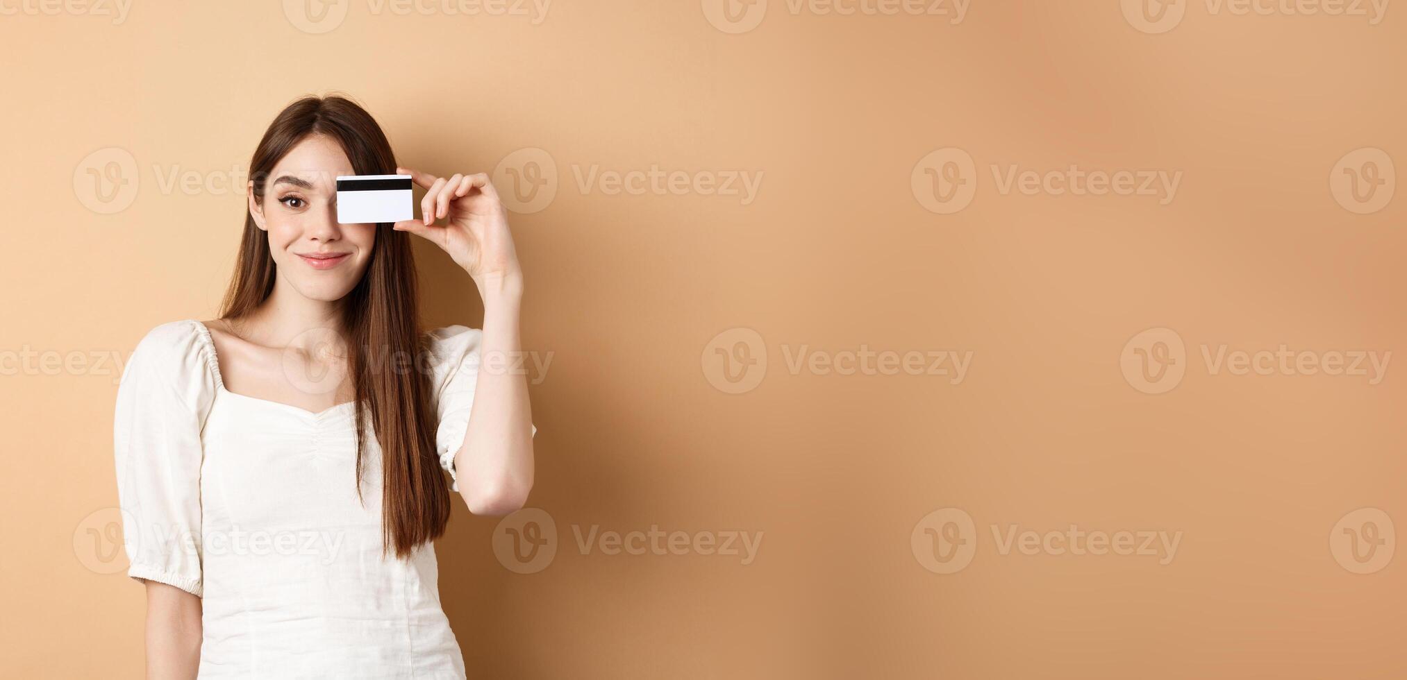 Smiling pretty girl showing plastic credit card over eye and looking satisfied, standing on beige background. Concept of shopping photo
