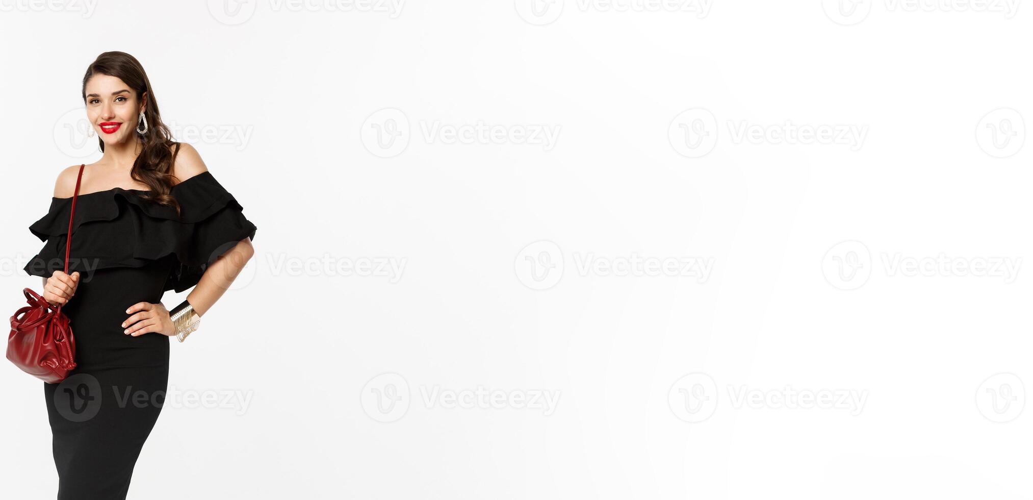 Beauty and fashion concept. Full length of elegant young woman going shopping in black dress, heels and purse, looking confident, standing over white background photo