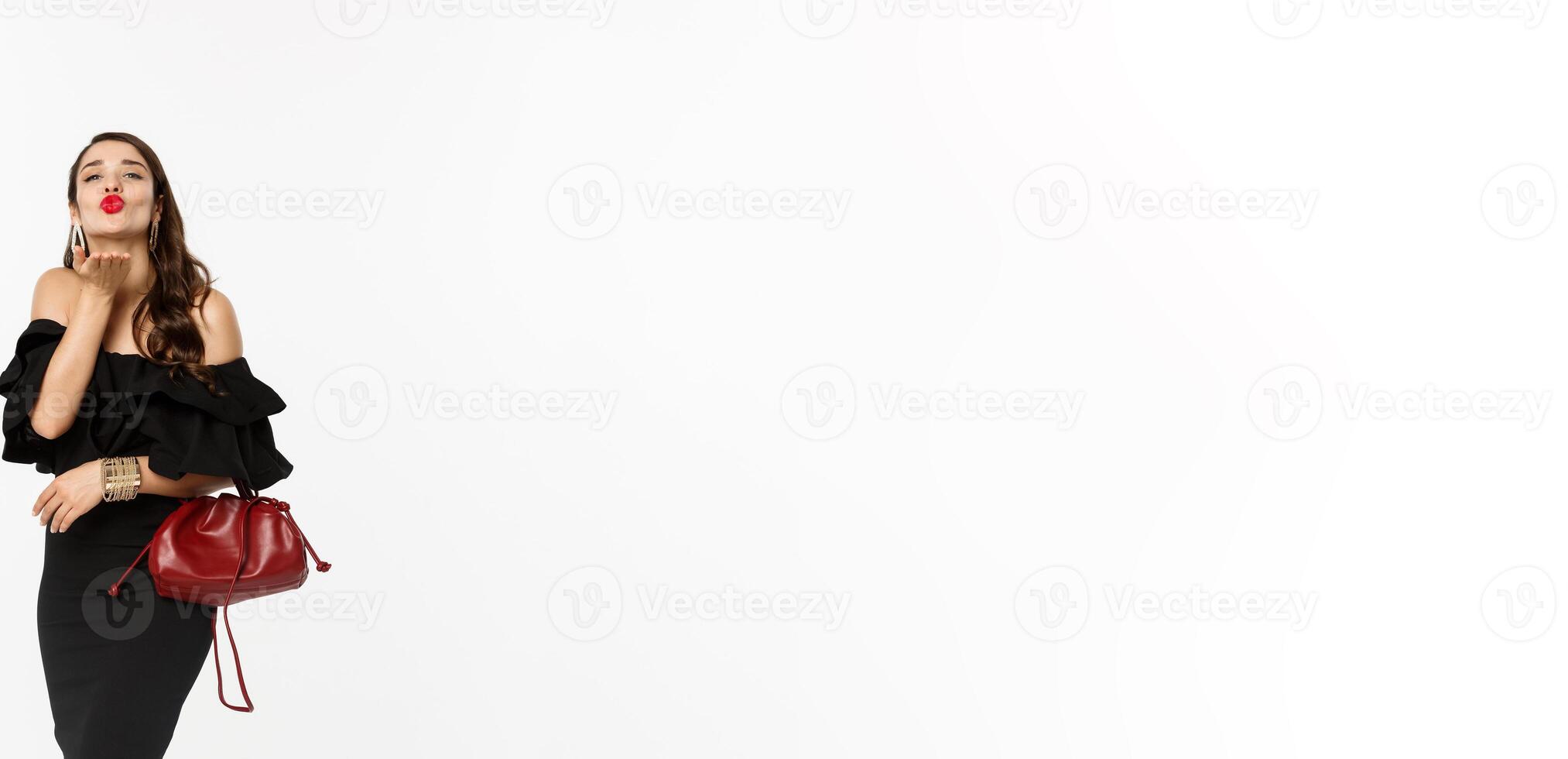 Beauty and fashion concept. Full length of glamour woman with red lipstick and makeup, wearing black dress with purse, sending air kiss at camera, white background photo