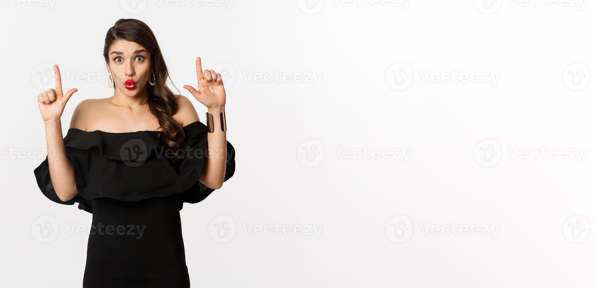 Fashion and beauty. Surprised woman showing advertisement on top, pointing fingers up and saying wow amazed, standing over white background photo