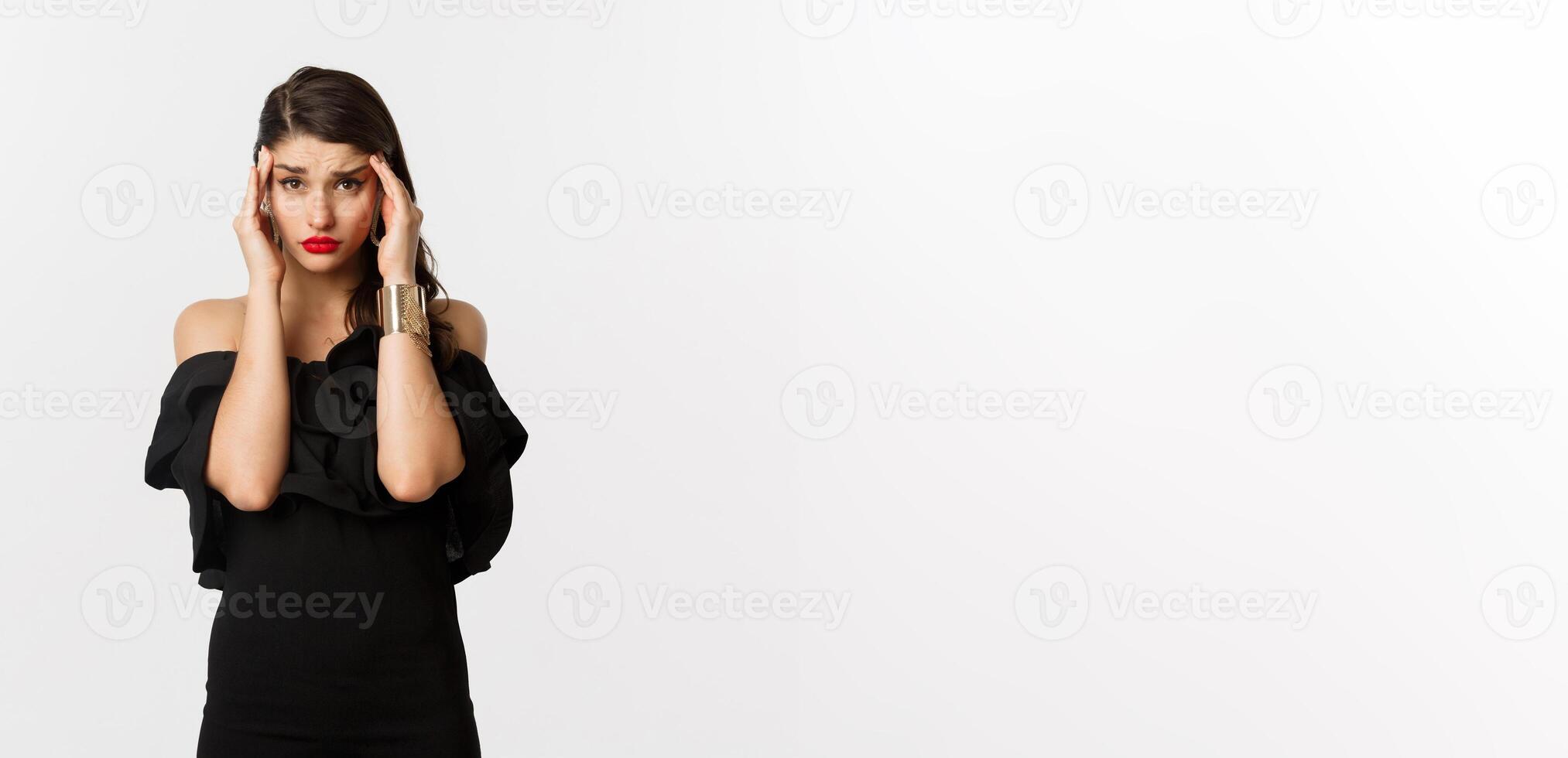 Fashion and beauty. Young modern woman in glamour dress, jewelry and makeup, touching head and looking exhausted, feeling dizzy, standing over white background photo