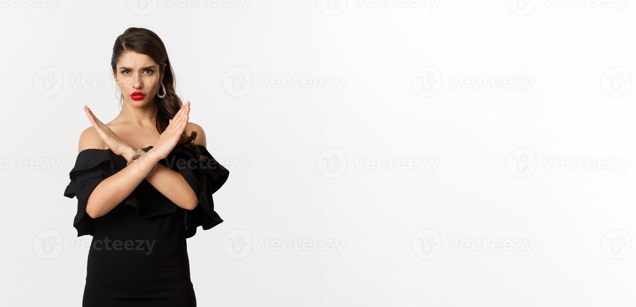 Fashion and beauty. Serious and confident female model in black dress, showing cross sign and frowning, stop gesture, telling no, standing over white background photo