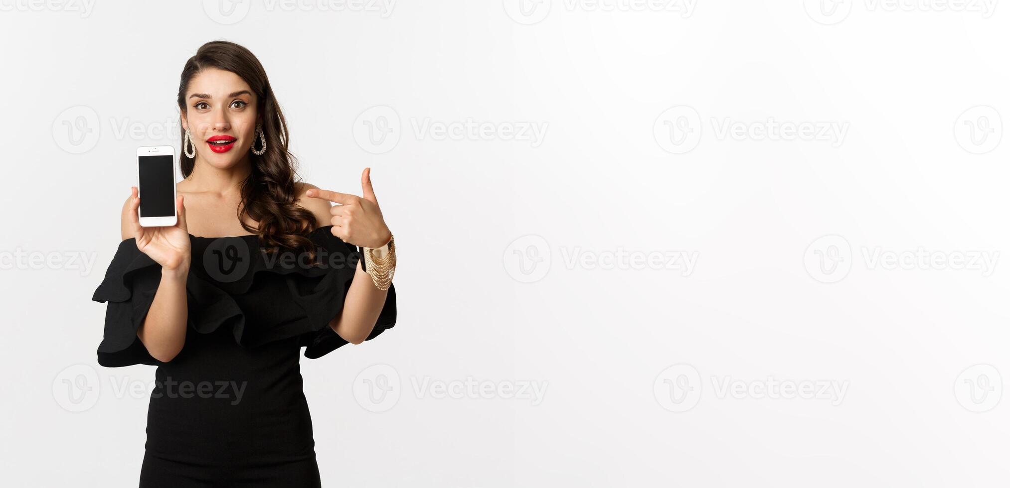 Online shopping concept. Fashionable woman in black dress pointing finger at smartphone screen, showing application, standing over white background photo