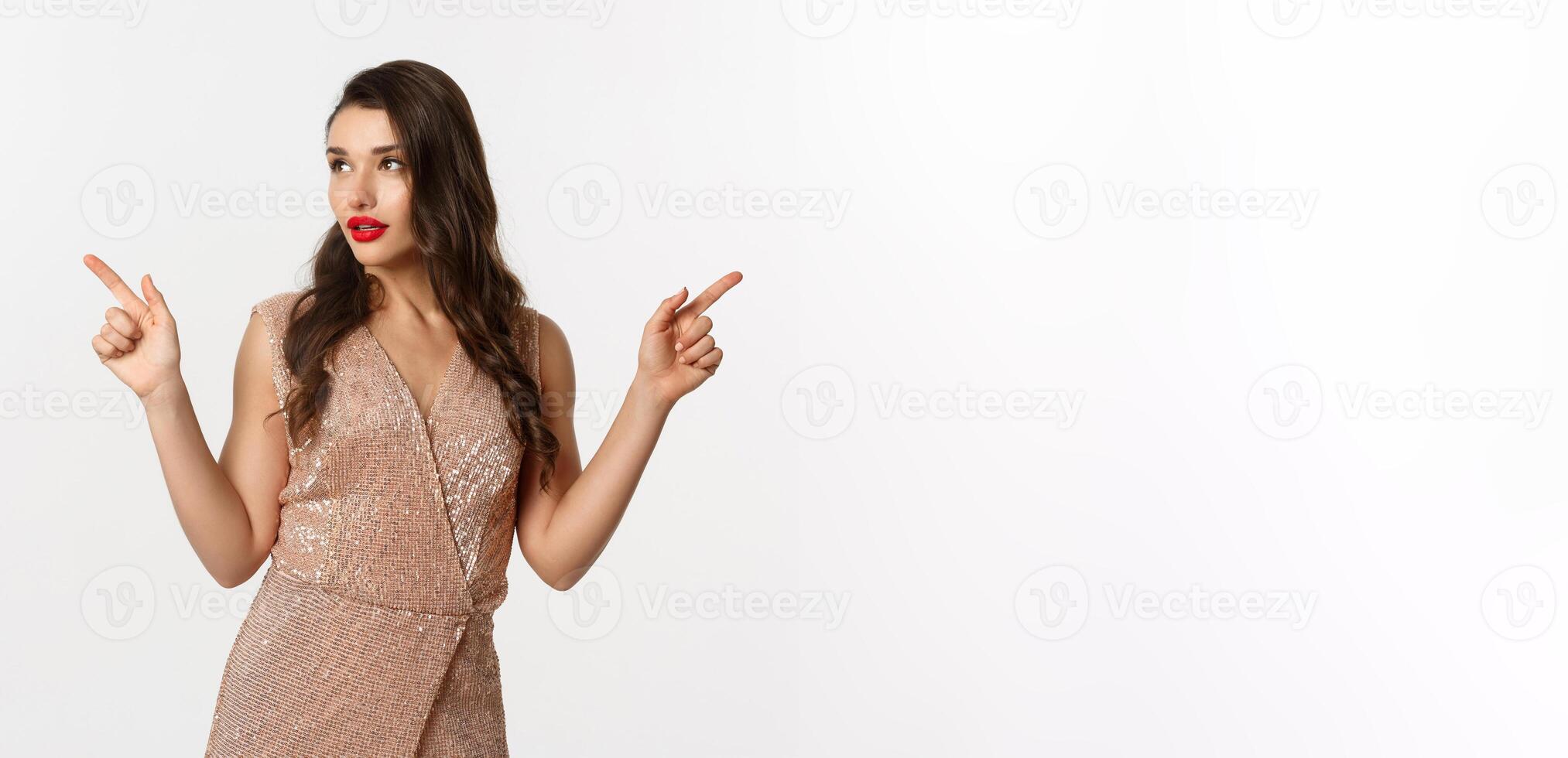 Luxury woman with red lips, wearing elegant evening dress, pointing fingers sideways and looking left at logo, showing christmas advertisement, stading over white background photo