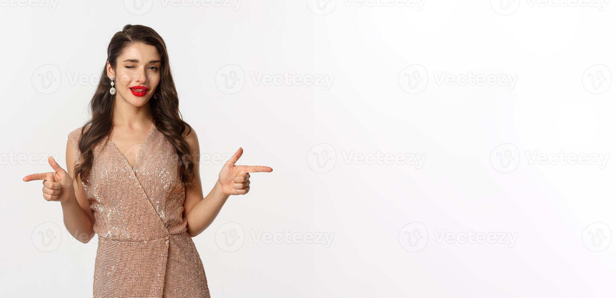 Concept of celebration, holidays and party. Beautiful and flirty woman with red lips, wearing glamour dress and winking, pointing fingers at camera, standing over white background photo