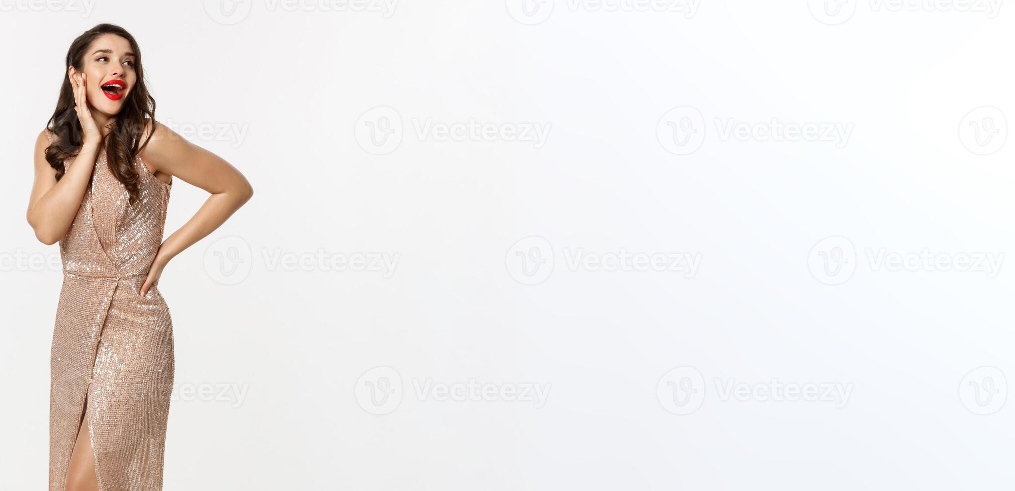 Party and celebration concept. Full-length of elegant woman in evening dress, standing near Christmas presents and looking surprised, standing over white background photo