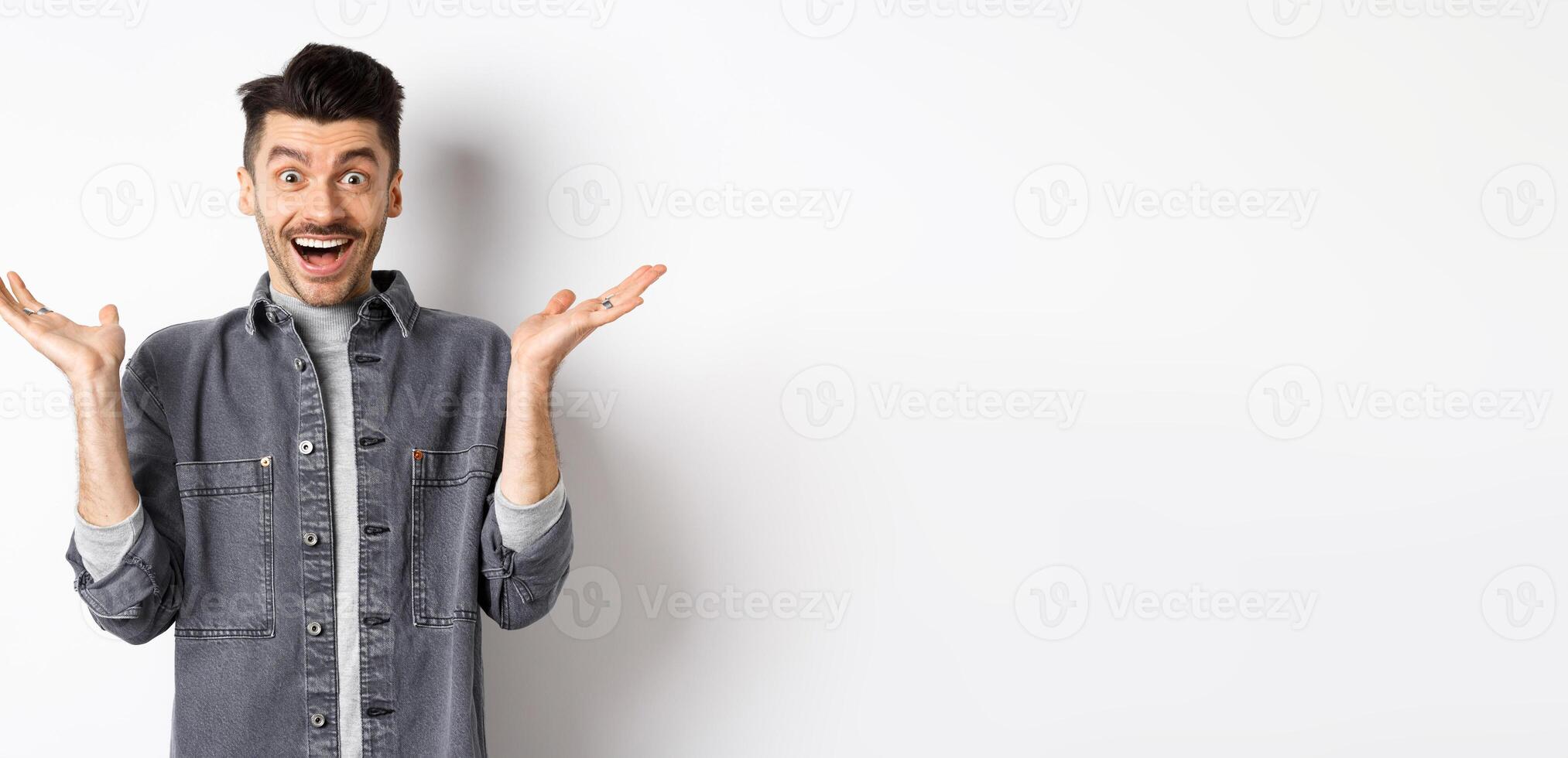 Excited young man cheering from good news, spread hands sideways and looking happy, smiling amazed, standing on white background photo