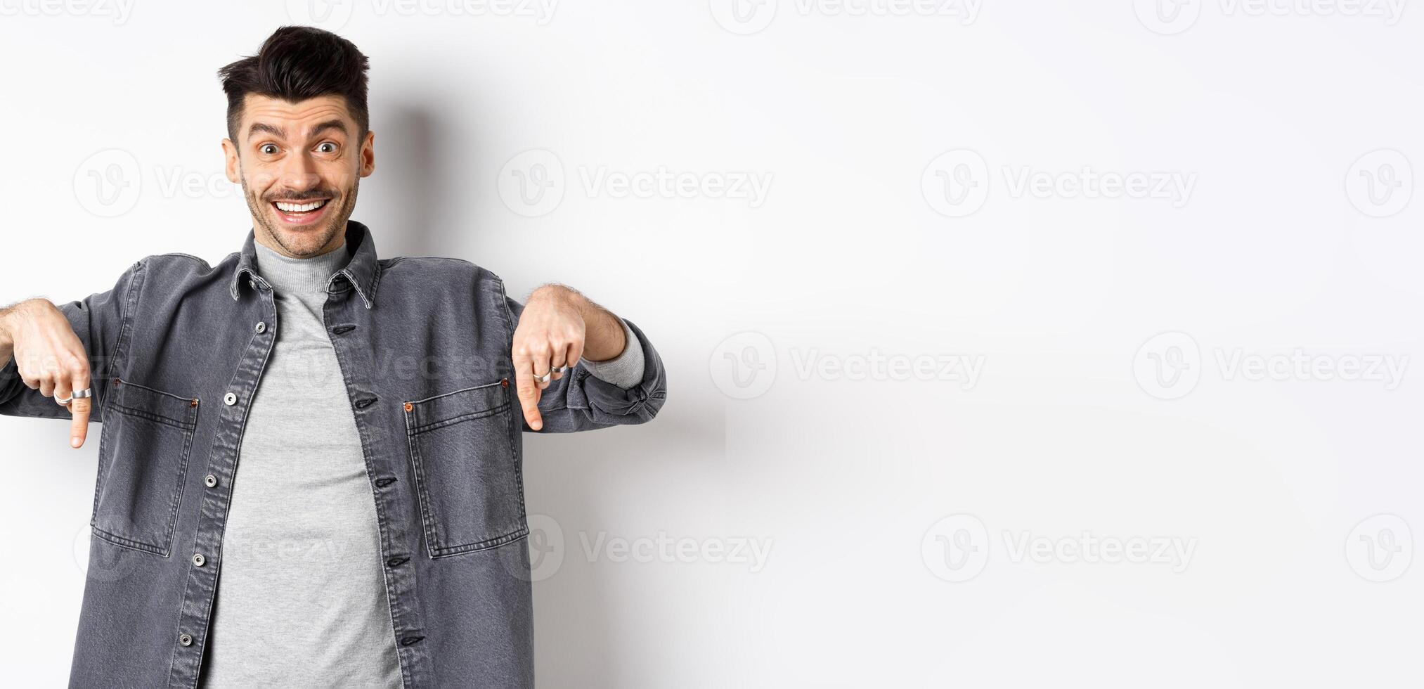 Excited smiling male model pointing fingers down, showing logo with happy face, demonstrate cool promo, standing on white background photo