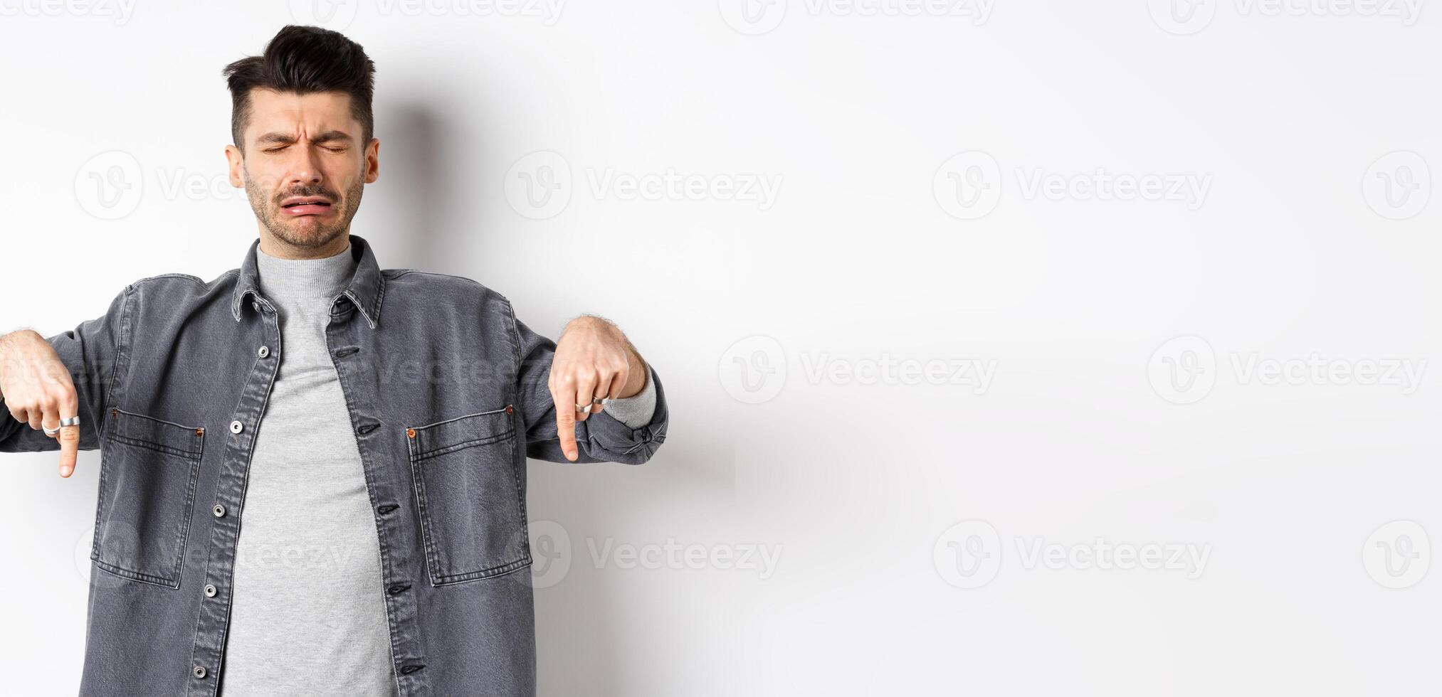 Sad crying man whining, pointing fingers down disappointed, showing bad news, standing upset on white background photo