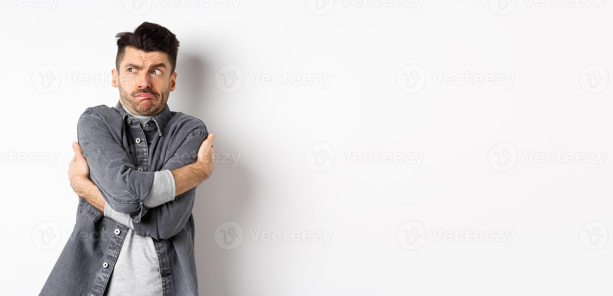 Displeased young man feeling discomfort, hugging himself and staring left at something embarrassing or exposing, standing uncomfortable on white background photo