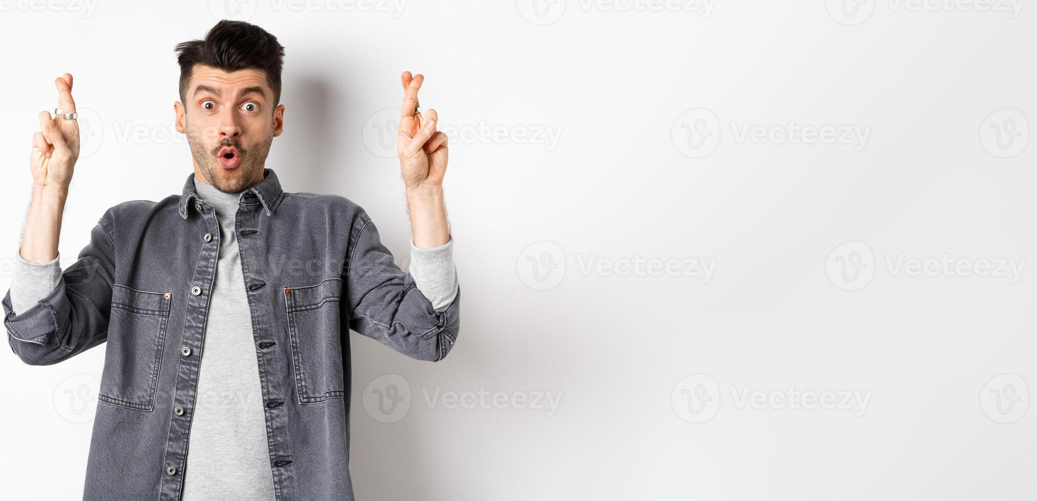 Excited young man cross fingers for good luck and gasping, holding breath as waiting important news, begging or praying, standing on white background photo