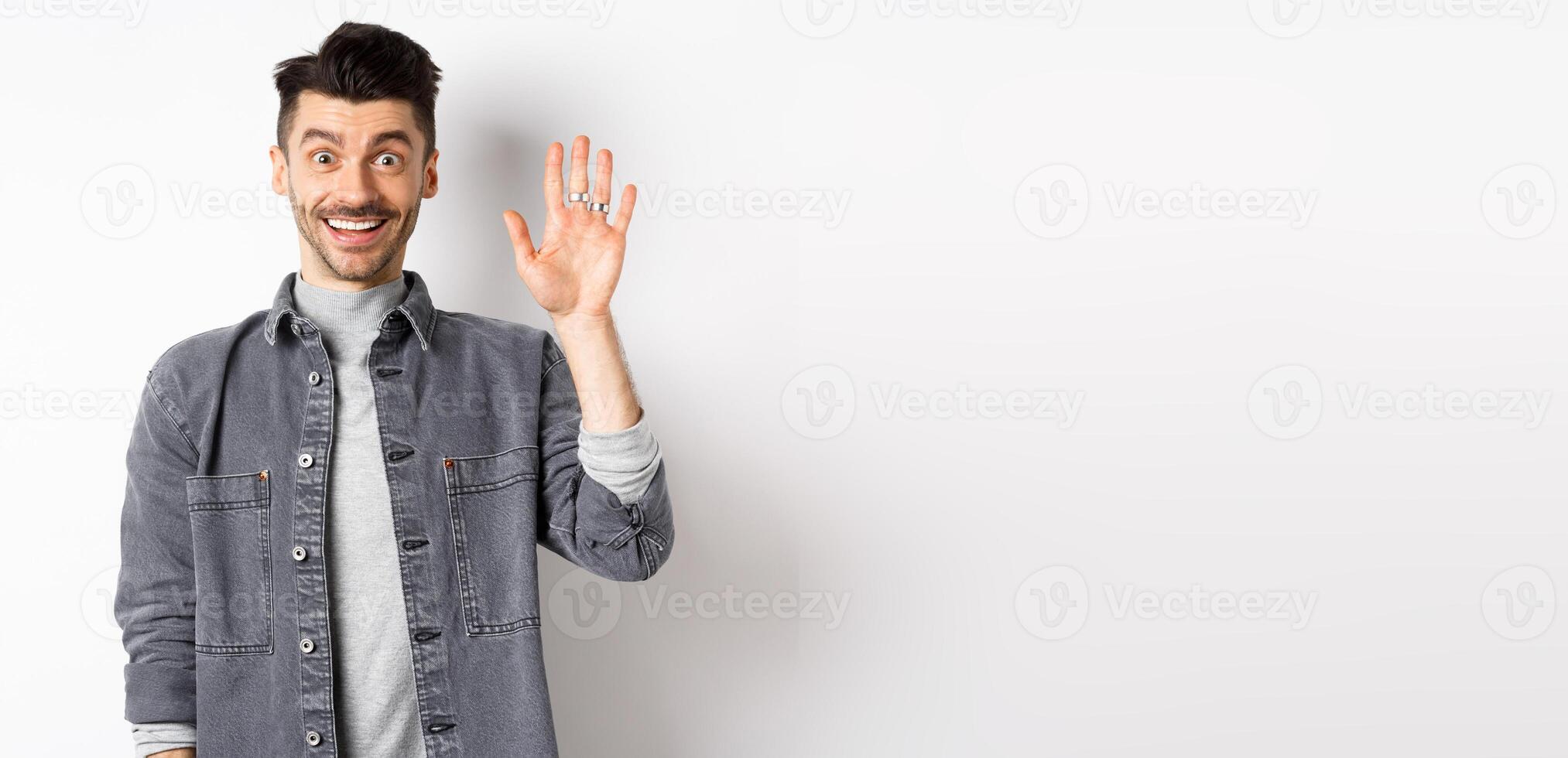 Friendly stylish man saying hello and waiving hand, smiling cheerful, greeting you with hi gesture, standing on white background photo
