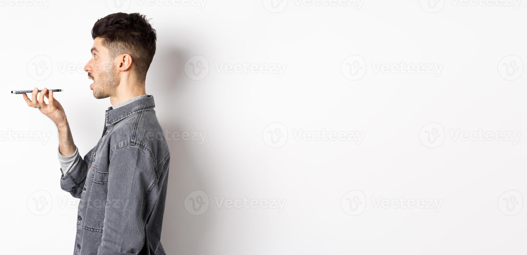 Profile of young man speaking at smartphone, translate with voice-translator app, talking into mobile phone speakerphone, standing on white background photo
