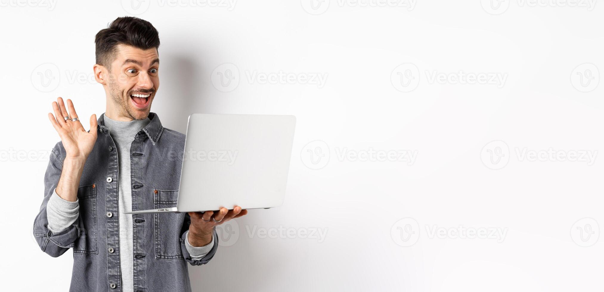 Excited man waiving hand at laptop, chatting on computer and smiling friendly, standing on white background photo