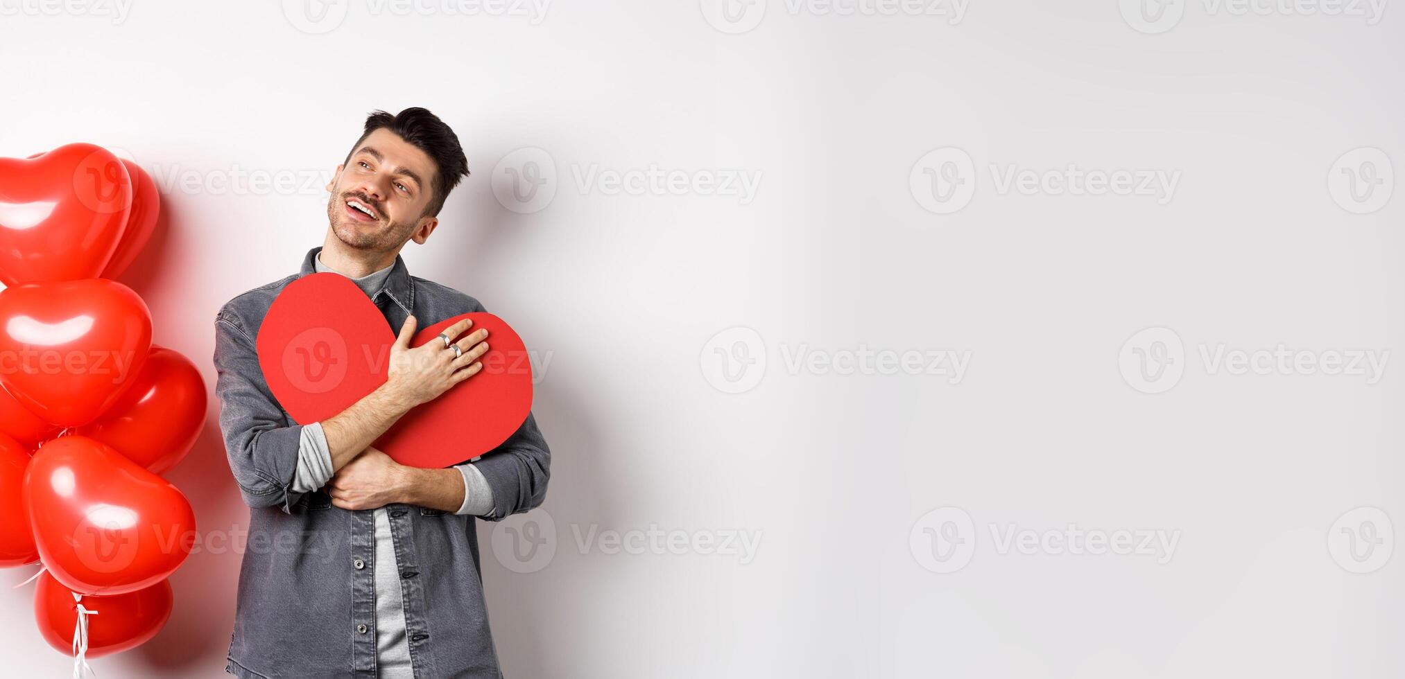 Romantic man hugging big red heart cutout and looking away dreamy, thinking of girlfriend and valentines day, imaging lover, standing on white background photo