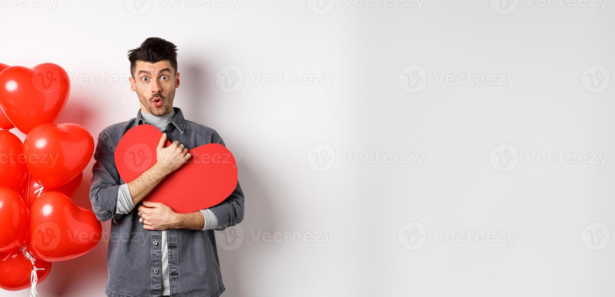Surprised man holding Valentines heart card and saying wow, looking amazed at camera, receive secret confession on lovers day, standing near romantic balloons on white background photo