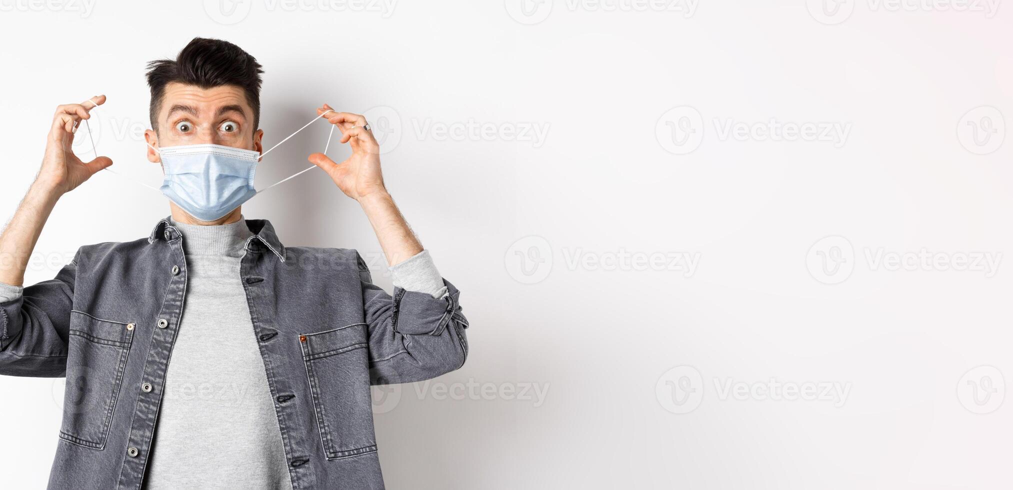 Health, covid and pandemic concept. Funny guy playing with medical mask on face, showing faces, standing against white background photo