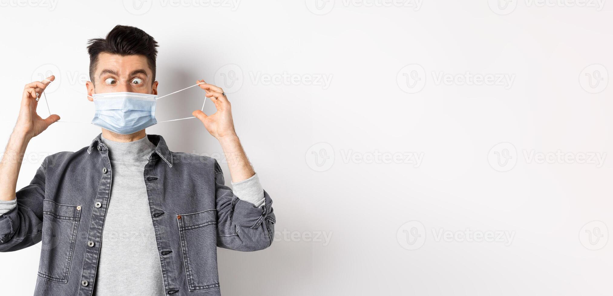 Health, covid and pandemic concept. Funny man squinting eyes at medical mask, playing with it, standing on white background photo