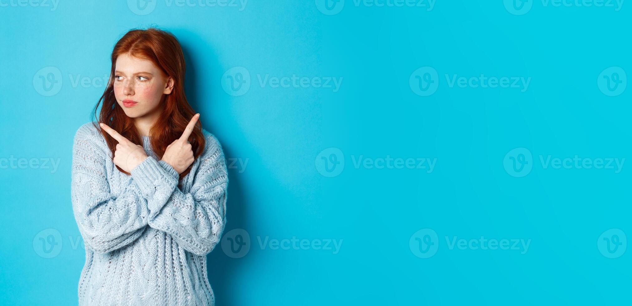 Indecisive redhead teenage girl making decision, pointing fingers sideways and looking left doubtful, standing in sweater against blue background photo
