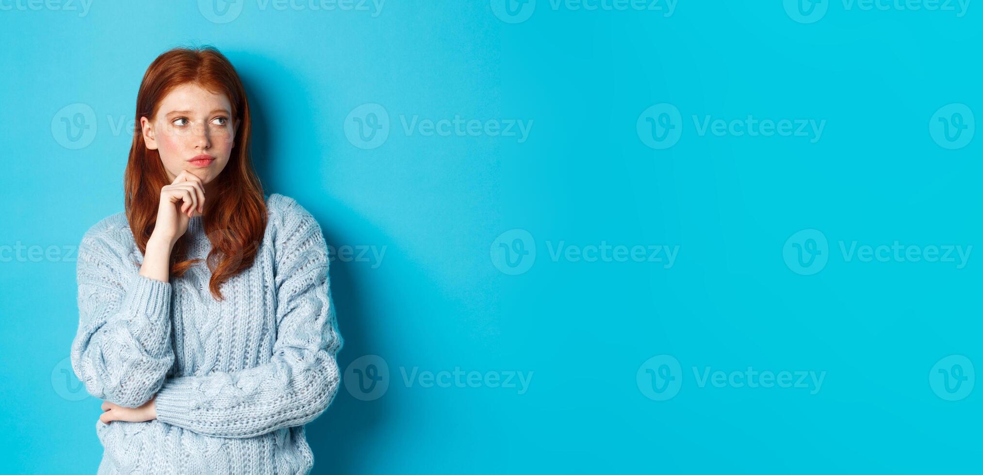Thoughtful and upset redhead girl looking right, pondering solution, standing in sweater against blue background photo