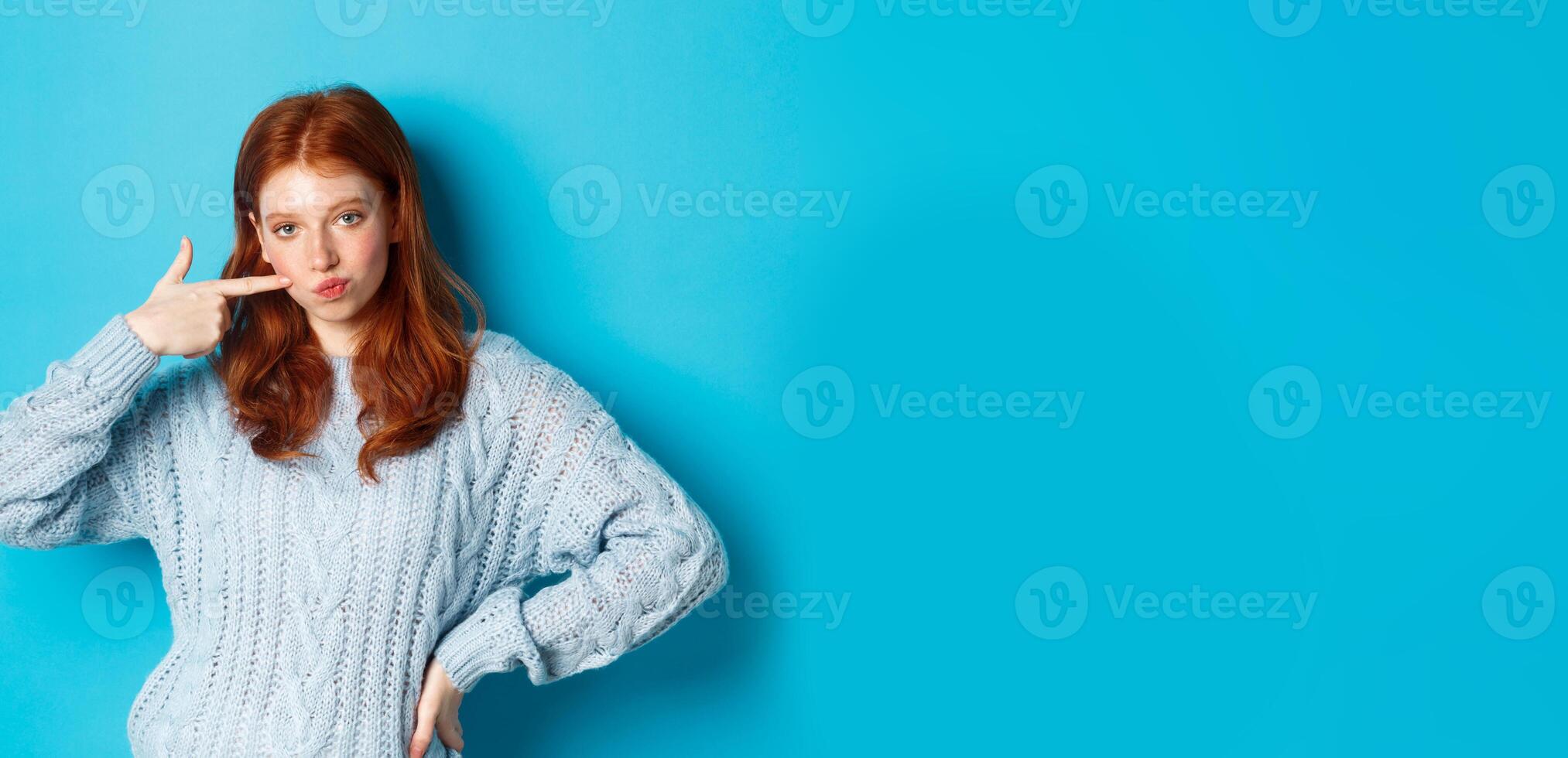 Cute redhead girl in sweater poking her cheek, staring at camera sassy, standing over blue background photo