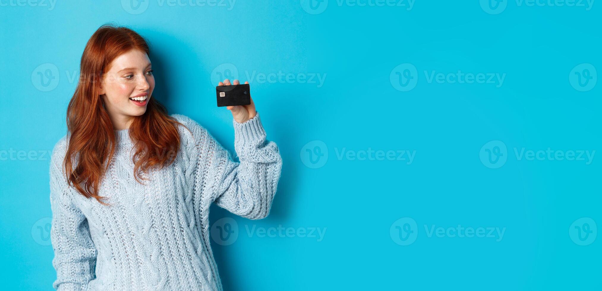 Cute redhead girl in sweater showing credit card, smiling at camera, standing over blue background photo