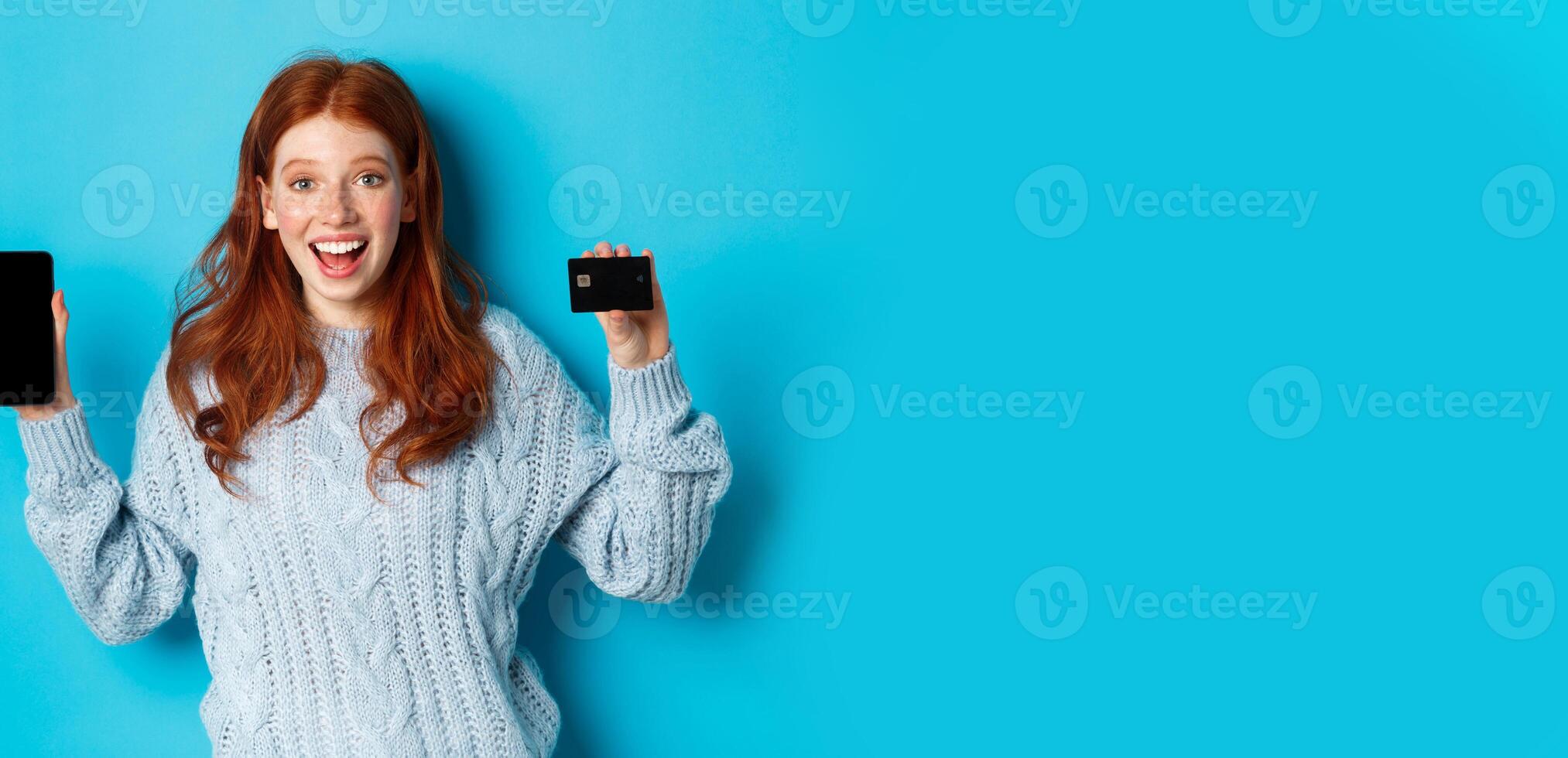Excited redhead girl showing mobile phone screen and credit card, demonstrating online store or application, standing over blue background photo