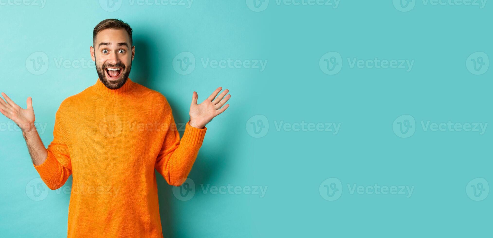 Excited man react to promo offer, spread hands sideways and rejoicing, standing against turquoise background in orange sweater photo