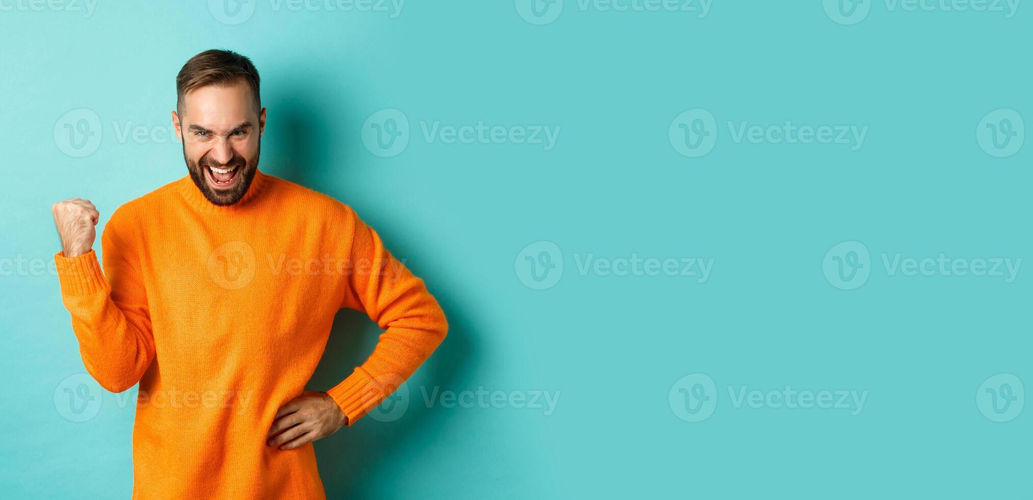 Image of satisfied young man feeling winner, fist pump and saying yes with satisfaction, achieve goal, triumphing while standing over light blue background photo