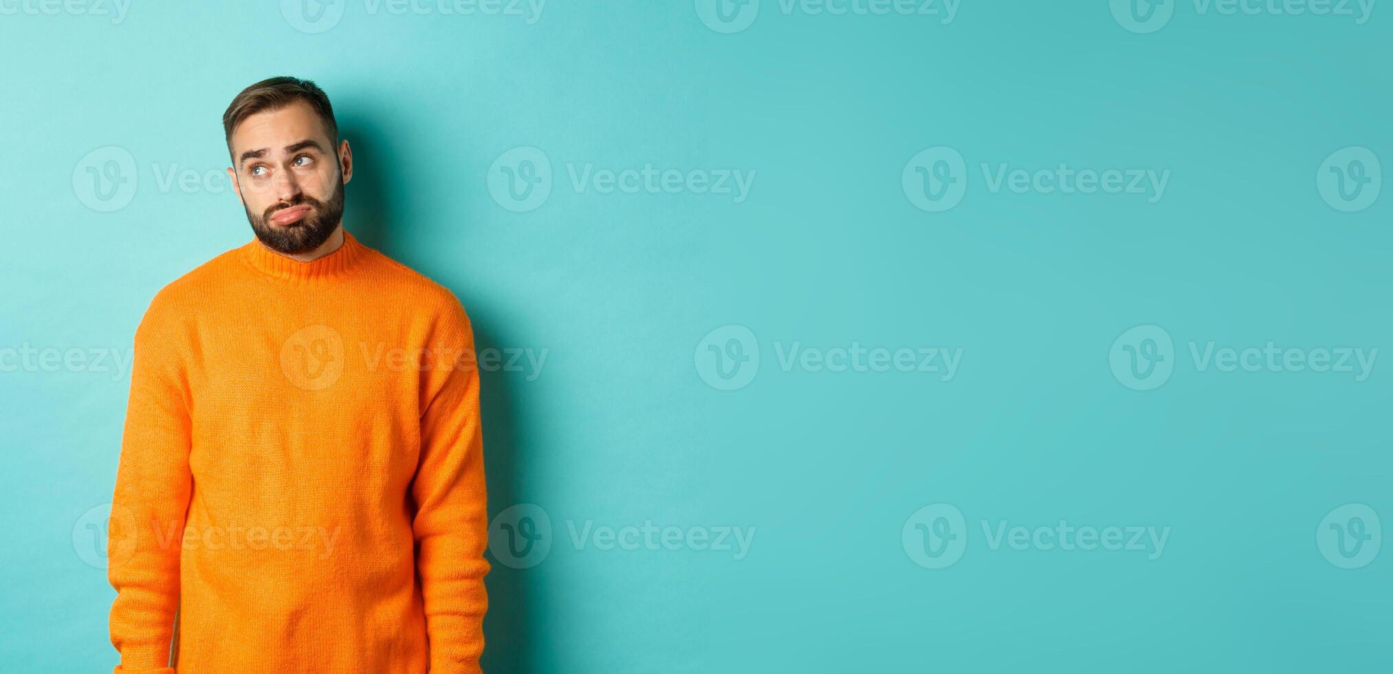 Sad and gloomy man sulking, looking bored at upper left corner, standing in orange sweater against light blue background photo