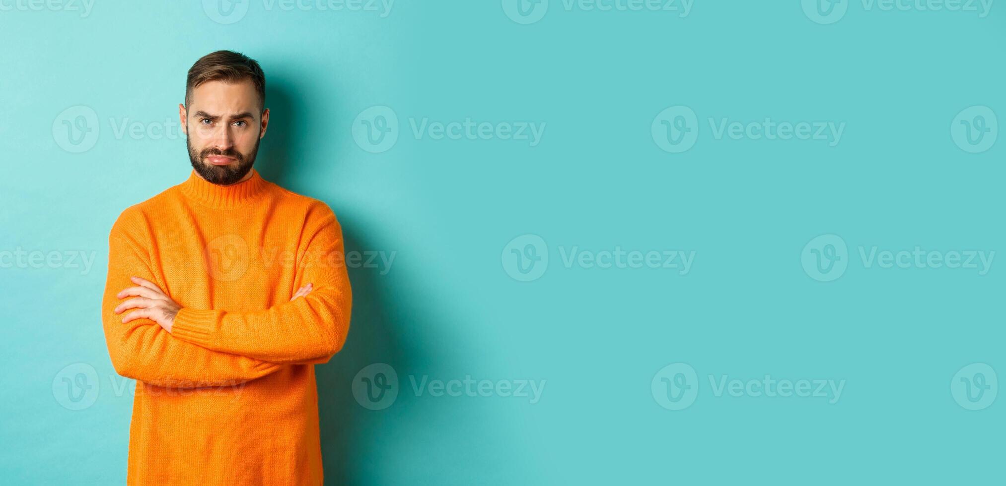 Silly sad guy with beard, sulking and looking offended, cross arms on chest and frowning upset, standing over light blue background photo