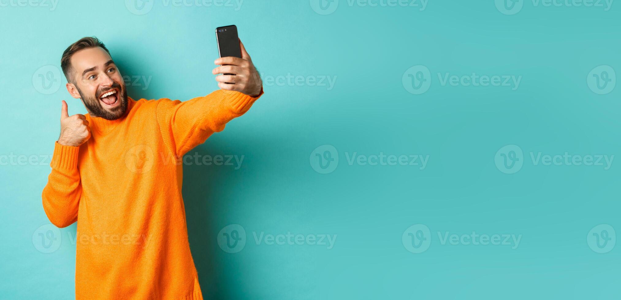 Handsome young man taking selfie on mobile phone, showing thumbs up at smartphone camera, recording vlogg, standing over light blue background photo